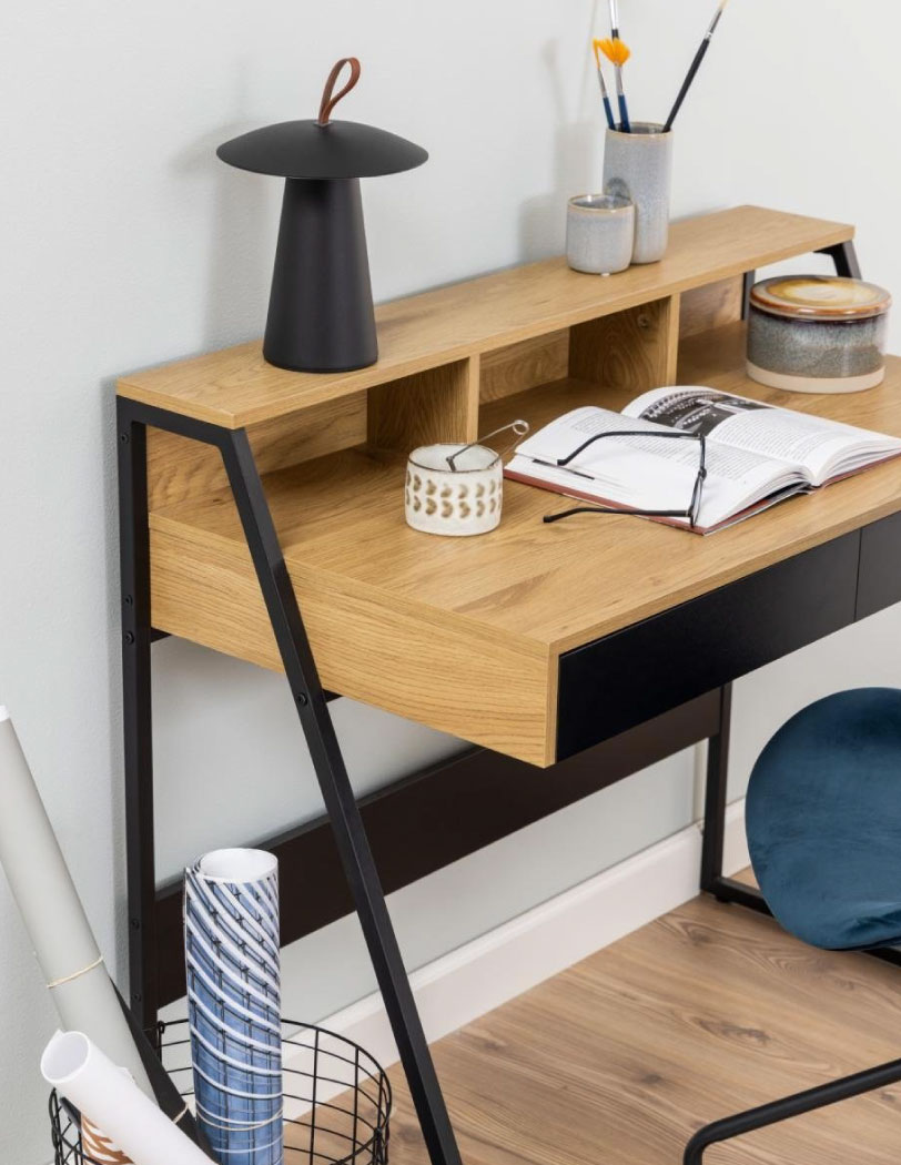 Fragment drewnianego biurka z czarnymi szufladami i nadstawką. Na biurku ksiażki, lampka oraz inne przybory do pracy.