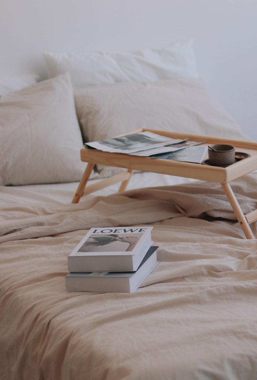 Zbliżenie na łóżko, na którym leżą książki oraz drewniana taca z magazynami oraz kubkiem.