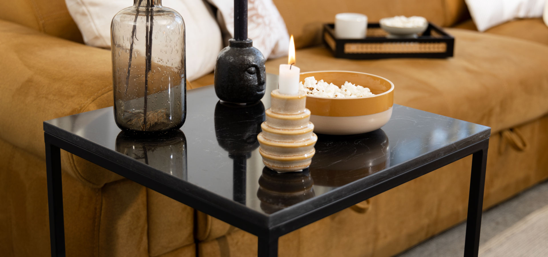 Czarny stolik z marmurowym blatem, na którym stoi wazon, dwa świeczniki oraz miseczka z popcornem. w tle musztardowa kanapa