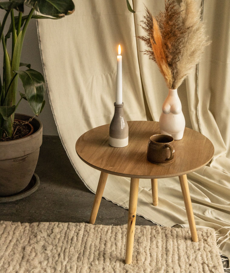 Okrągły stolik kawowy z imitacji drewna, na którym stoi wazon w kształcie kobiety z suszoną trawą, świecznik i kamionkowy kubek. w tle duża donica z rośliną