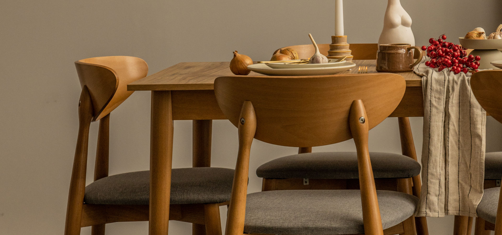 Drewniany stół oraz krzesła w jadalni. Krzesła z zaokrąglonym oparciem oraz tapicerowanym, szarym siedziskiem. Na stole ustawione naczynia, wazon oraz świeczka.