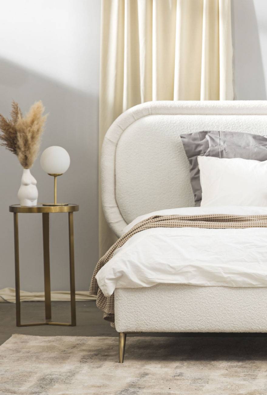 Białe łóżko w tkaninie boucle z dużym zagłówkiem w organicznym kształcie. Po lewej stronie łóżka wysoki stolik pomocniczny w złotym kolorze