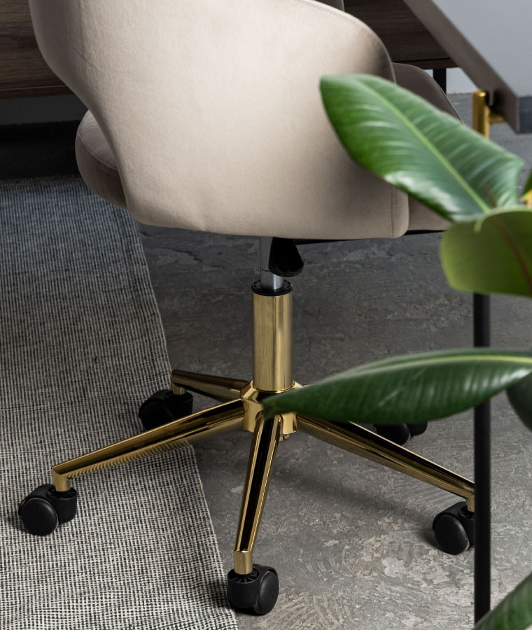 Beżowe krzesło na złotych nogach z wycięciem w oparciu, stojące przy czarnym biurku na cienkich, metalowych nogach.