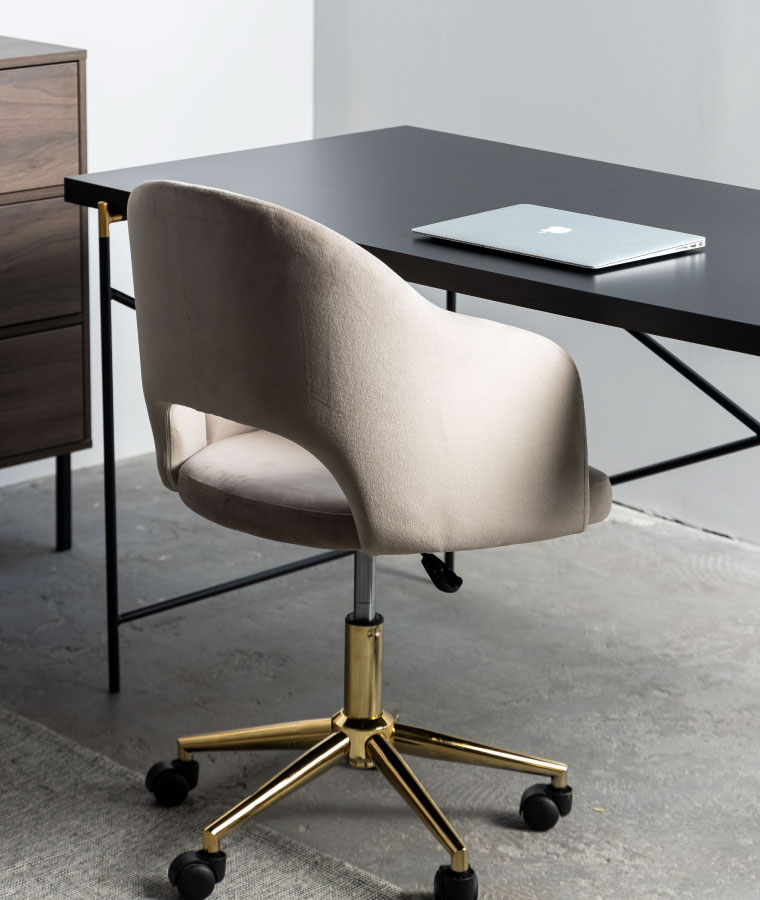 Beżowe krzesło na złotych nogach z wycięciem w oparciu, stojące przy czarnym biurku na cienkich, metalowych nogach.