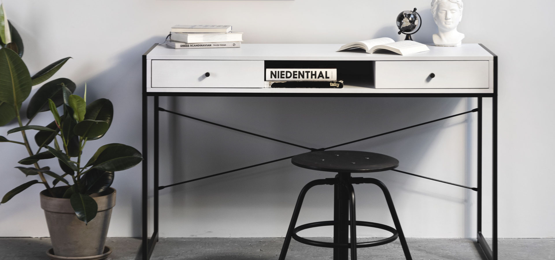 Białe biurko z dwoma szufladami stojące na metalowej konstrukcji. Obok biurka widoczna duża roślina w betonowej donicy. Przed biurkiem stoi niski, czarny stołek w stylu industrialnym.