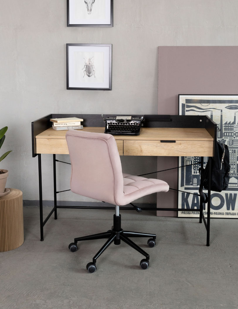 Różowe krzesło obrotowe stojące przy drewnianym biurku z czarną konstrukcją. Na biurku widoczna maszyna do pisania oraz książki. W tle obrazy zwierząt oraz ilustracja industrialnych Katowic