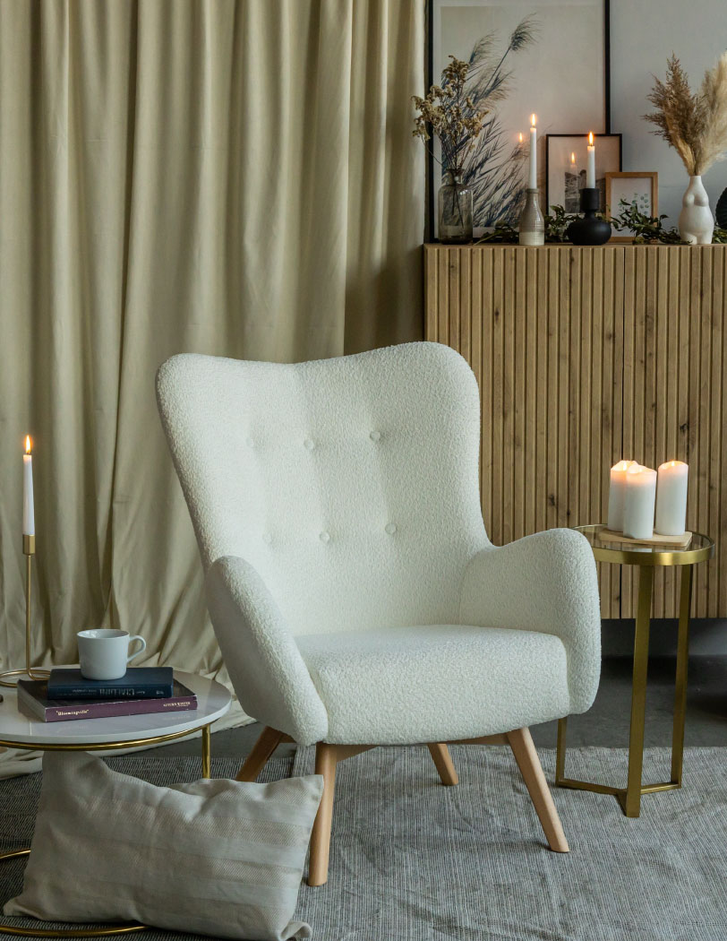 Beżowy fotel w stylu uszaka w tkaninie boucle na drewnianych nogach. W tle drewniana komoda oraz wysoki złoty stolik.