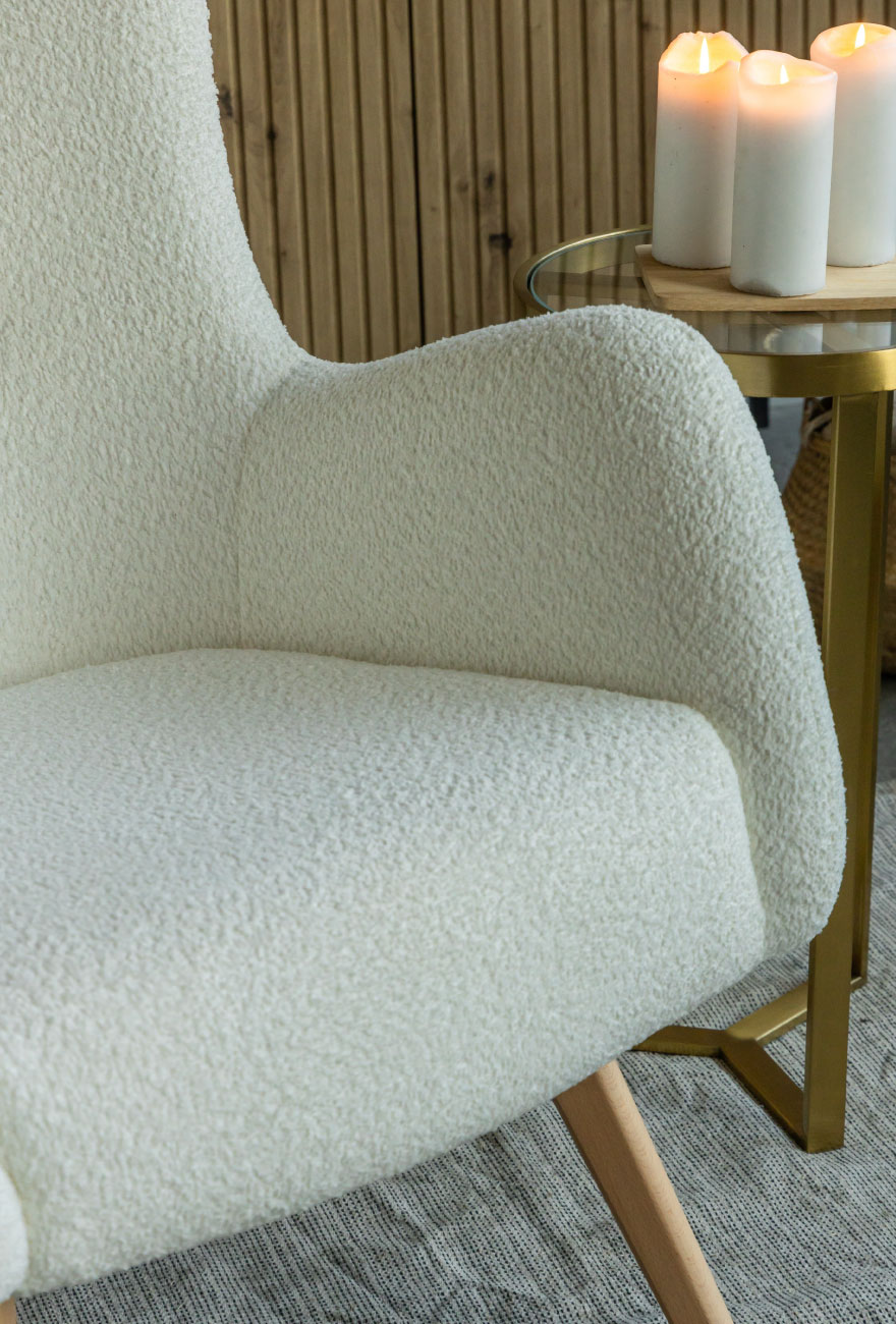 Beżowy fotel w stylu uszaka w tkaninie boucle na drewnianych nogach. W tle drewniana komoda oraz wysoki złoty stolik.