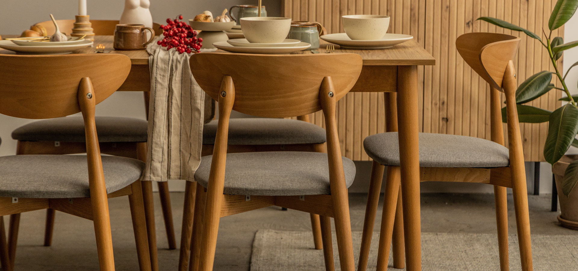 Drewniany stół z drewnianymi krzesłami o siwych siedziskach. Na stole zastawa w kolorze beżowym, w tle drewniana komoda oraz roślina.