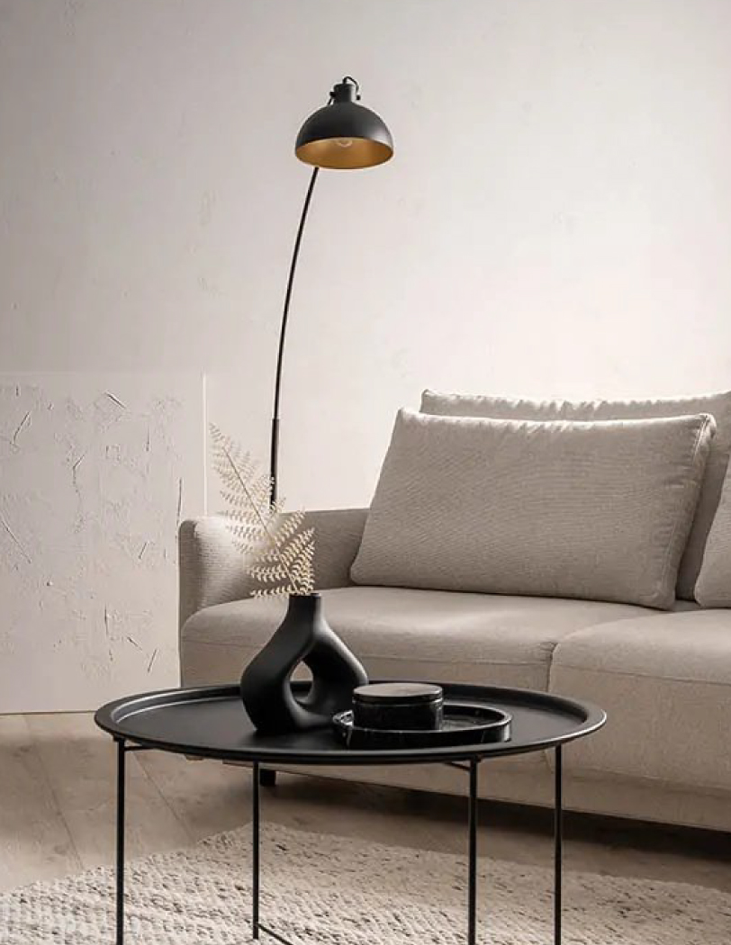 Lampa podłogowa w kolorze czarnym z wygiętym ramieniem sięgającym nad beżową kanapę. Przed kanapą czarny stolik kawowy z czarnymi dekoracjami.