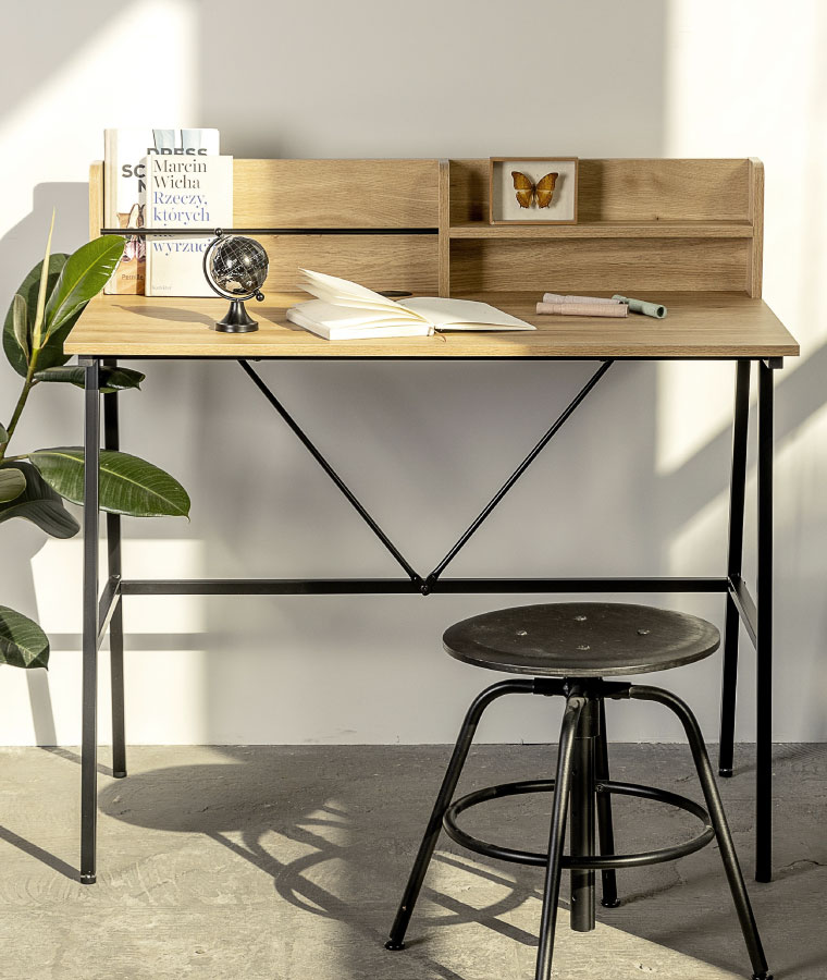Na zdjęciu widoczne biurko w jasnym, bukowym drewnie. Przed biurkiem stoi niski metalowy stołek w stylu industrialnym. Na biurku leżą między innymi książki i czarny globus.