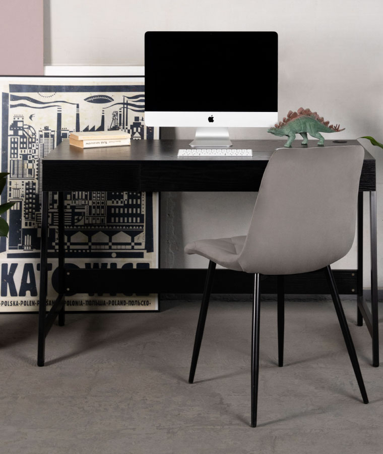 Na zdjęciu widoczne jest czarne biurko, na którym stoi komputer i leżą książki. Przed biurkiem stoi szare krzesło. W tle widoczne plakaty z wizerunkiem Katowic.