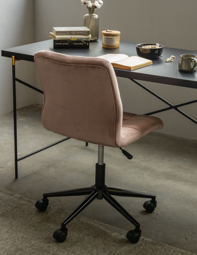 Na zdjęciu widoczny tył krzesła w odcieniu różowym, stojącego przy czarnym biurku. Na biurku książki, miseczka i kubek.