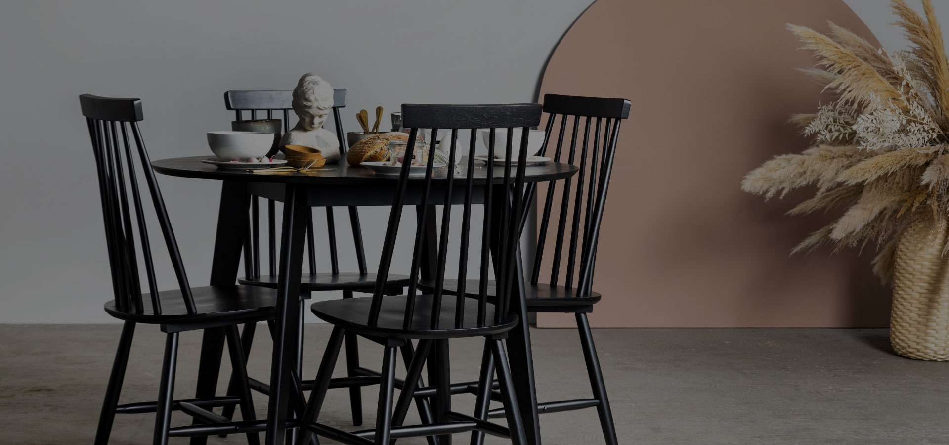 Czarny drewniany zestaw składający się ze stołu i krzeseł, na którym widać zastawę stołową. W tle kompozycja z suszonych kwiatów.