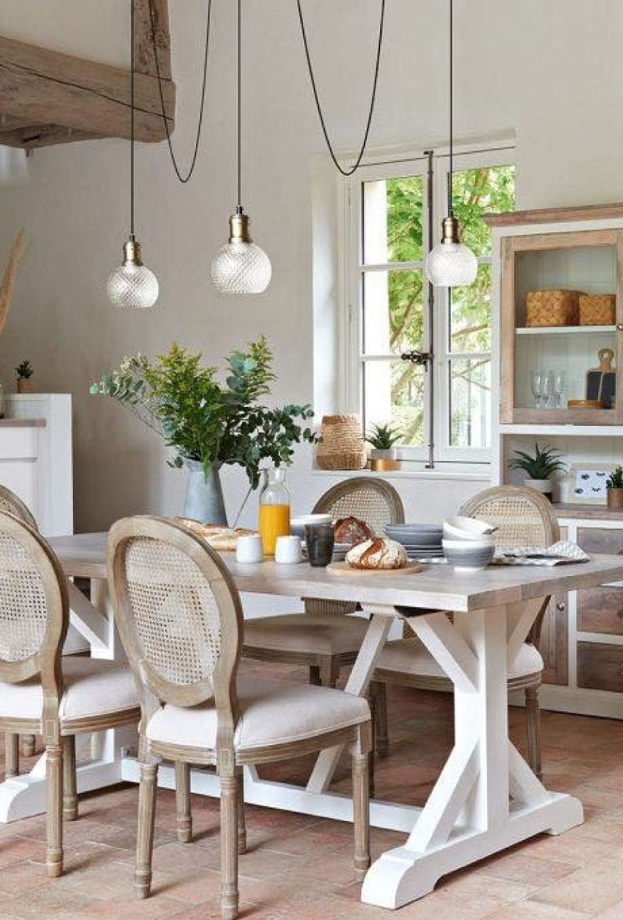 Wnętrze salonu w stylu modern farm house z dużym białym stołem i zdobnymi drewnianymi krzesłami. Nad stołem wisi lampa 3-punktowa z kryształowymi kloszami.