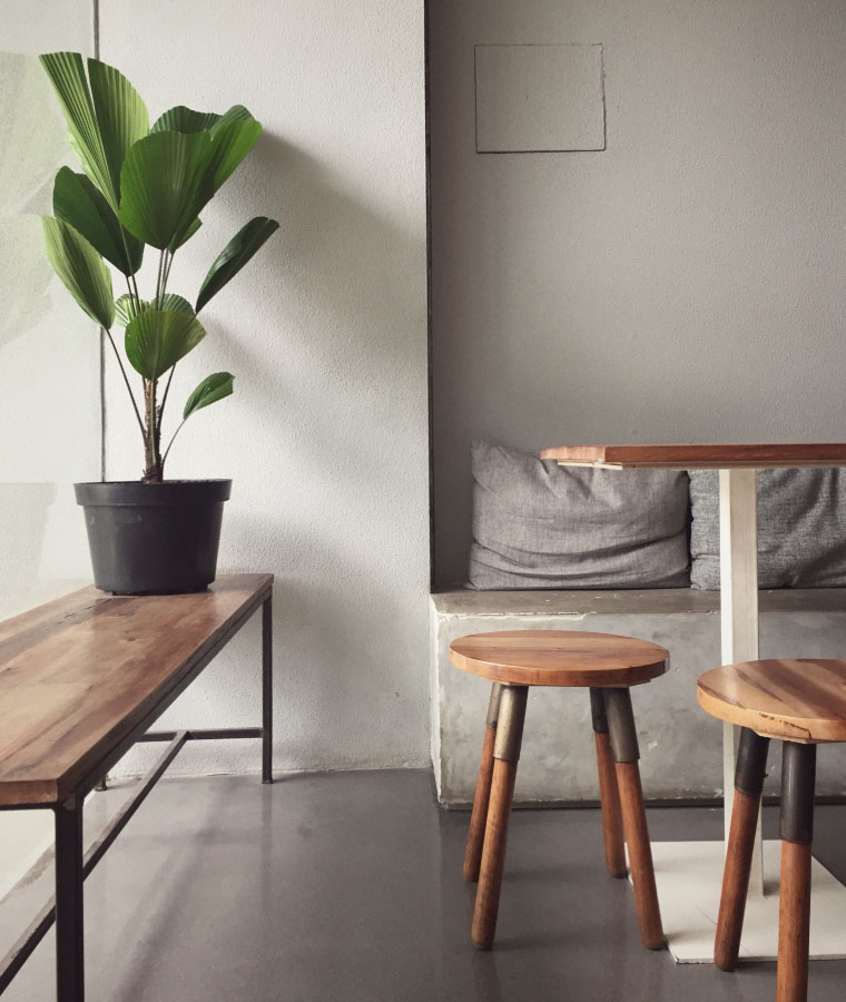 Wnętrze pokoju z betonową podłogą i ścianami. Z boku widoczny stół na metalowej nodze i z drewnianymi stołkami, po lewej stronie ławka w stylu loftowym ze stojącym kwiatem w czarnej donicy.
