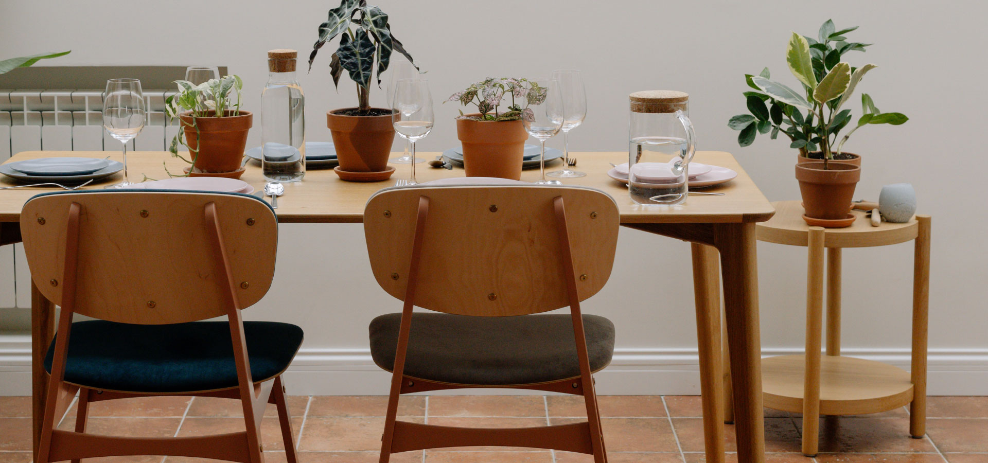 Wnętrze jadalni z widocznym drewnianym stołem i krzesłami z metalowej i drewnianej konstrukcji z tapicerowanym oparciem. Na stole widoczna zastawa i rośliny.