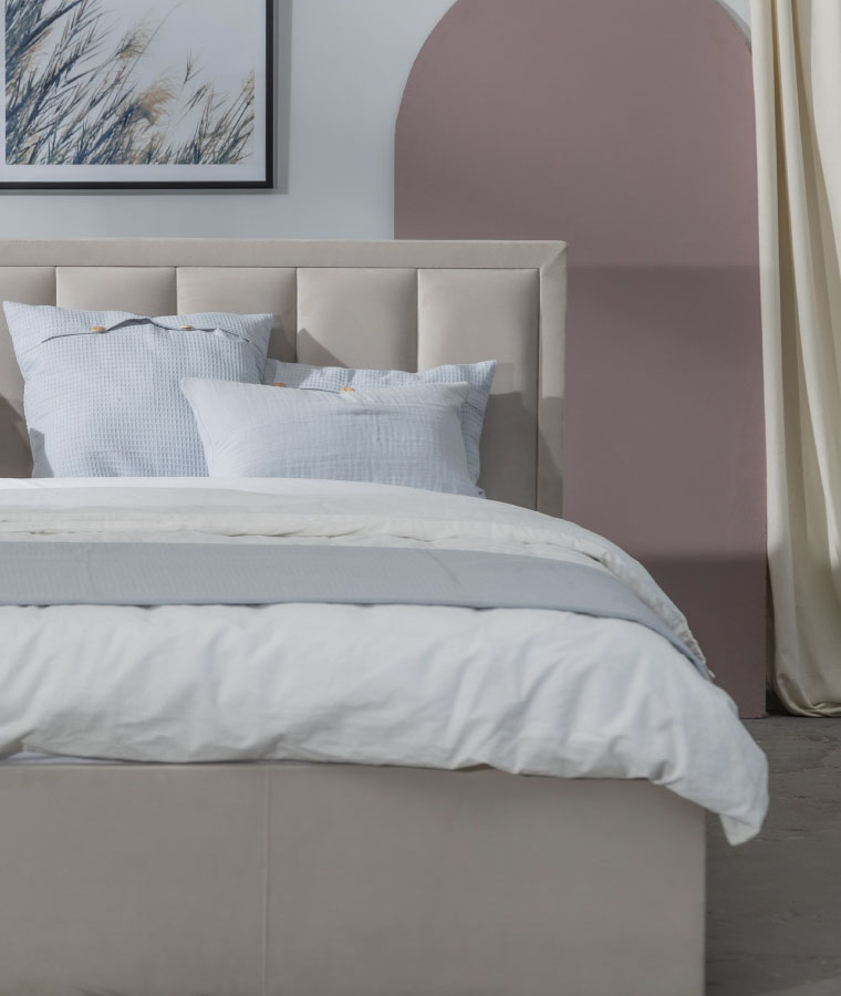 Jasnobeżowe łóżko z niskim zagłówkiem w pionowe pasy. Na łóżku widoczna pościel w kolorze białym i błękitne poduszki.