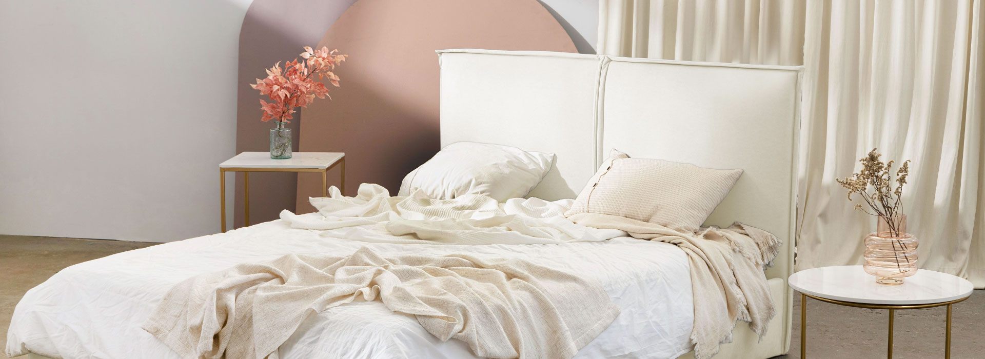 Łóżko tapicerowane w kolorze jasnobeżowym z gładkim zagłówkiem z pionowym przeszyciem na środku. Na łóżku widoczne poduszki oraz pościel, po bokach stoliki z marmurowymi blatami.