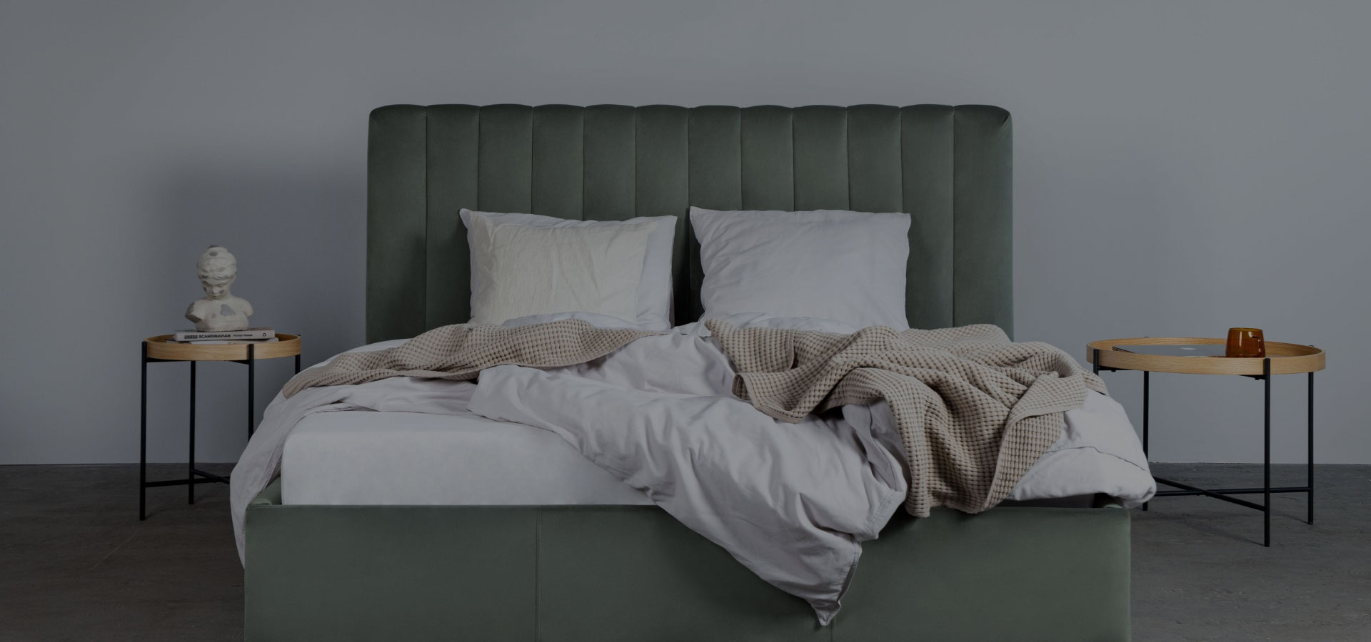 Pistacjowo-zielone łóżko z weluru z wysokim zagłówkiem z pionowymi przeszyciami. Na łózku widoczna pościel i poduszki. Po obu stronach łóżka stoliki na metalowych nogach z drewnianymi blatami