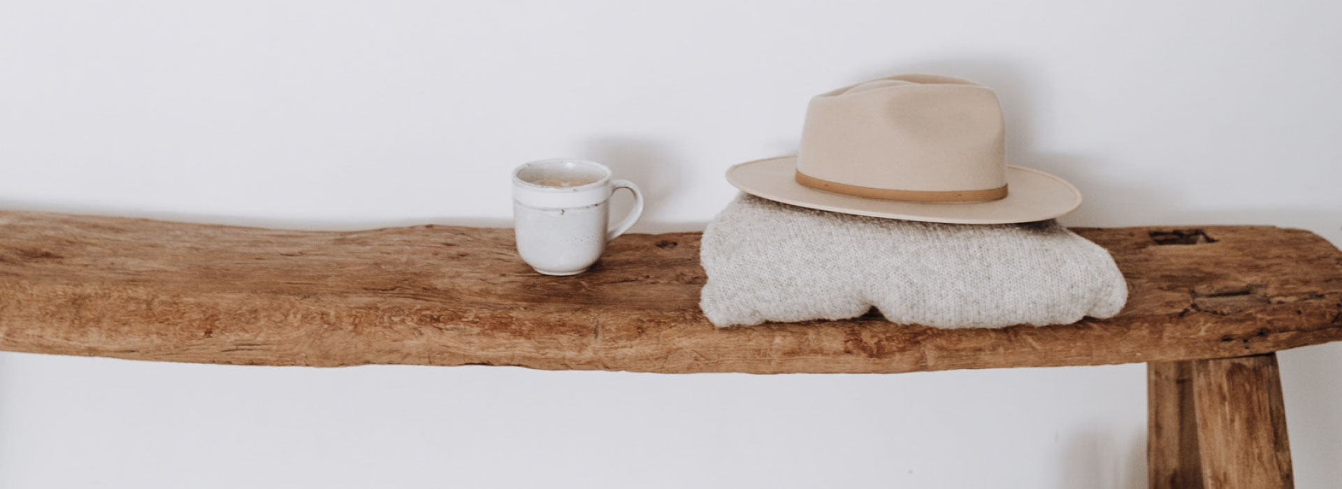 Drewniana ławka, na której stoi kubek z kawą oraz złożony sweter i kapelusz. Pod ławką stoją czarne buty i widoczny jest fragment dywanu.