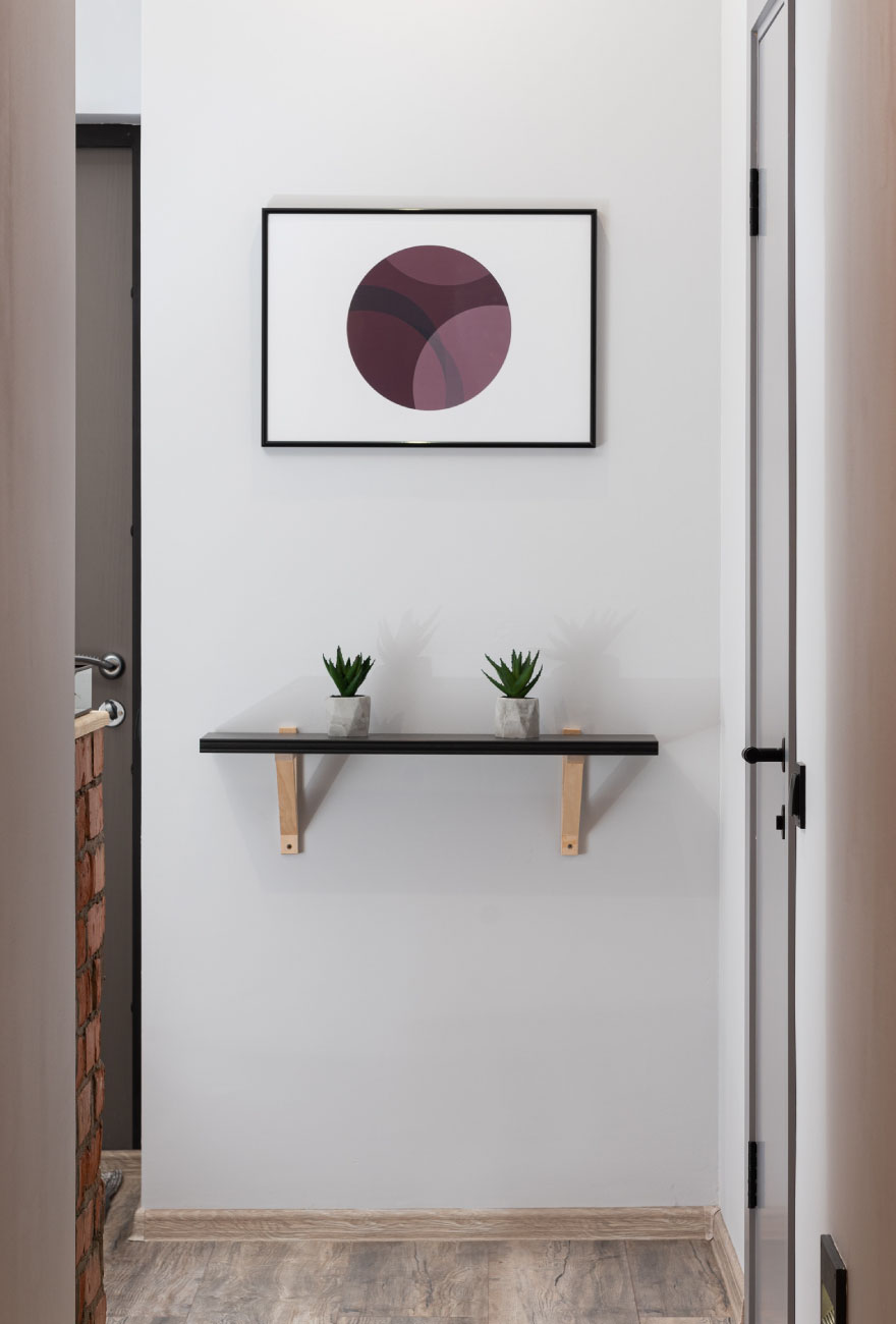 Fragment ściany, na której wisi drewniana półka z małymi dekoracjami ze sztucznych roślin. Nad półką widoczny plakat w czarnej ramie.