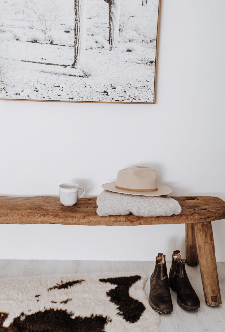 Fragment drewnianej ławki, na której stoi kubek oraz leży złożony sweter z kapeluszem. Pod ławką stoją czarne buty i widoczny jest fragment dywanu w łaty.