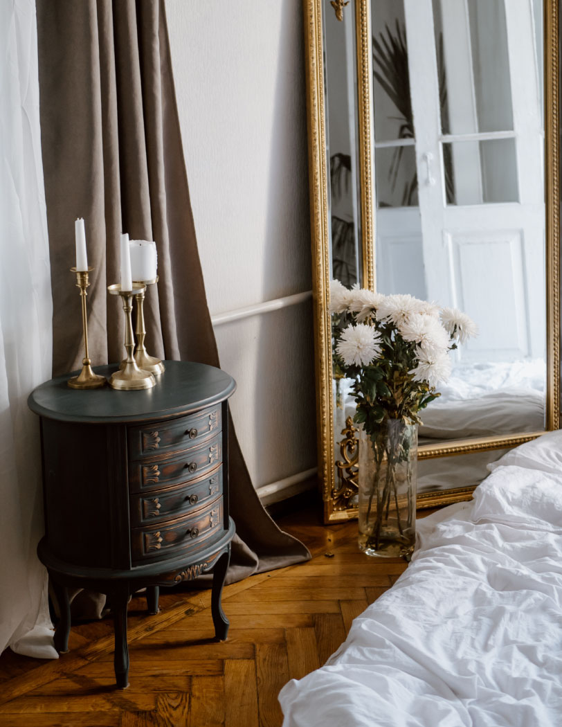 Wnętrze domu w starym stylu, w tle duże lustro w złotej ramie i wysoki szklany wazon z kwiatami. Po lewej stronie niska szafka o okrągłym kształcie z czterema szufladami i na giętych nogach.