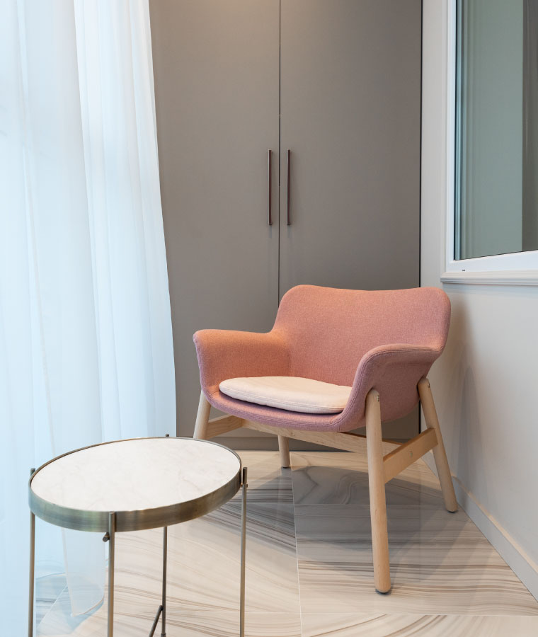 Różowy fotel na drewnianej konstrukcji z niskim stolikiem na złotych nogach i z marmurowym blatem. W tle szara szafa z czarnymi uchwytami.