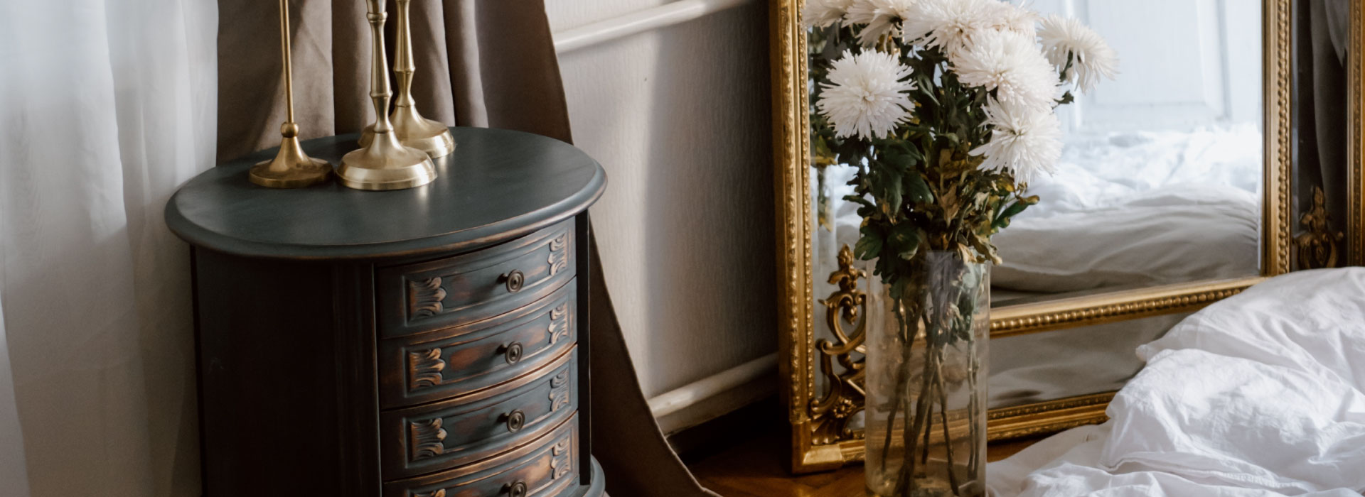 Zbliżenie na niską retro szarkę z szufladami o okrągłej formie. W tle lustro stojące na ziemi w złotej ramie oraz wysoki wazon z kwiatami.