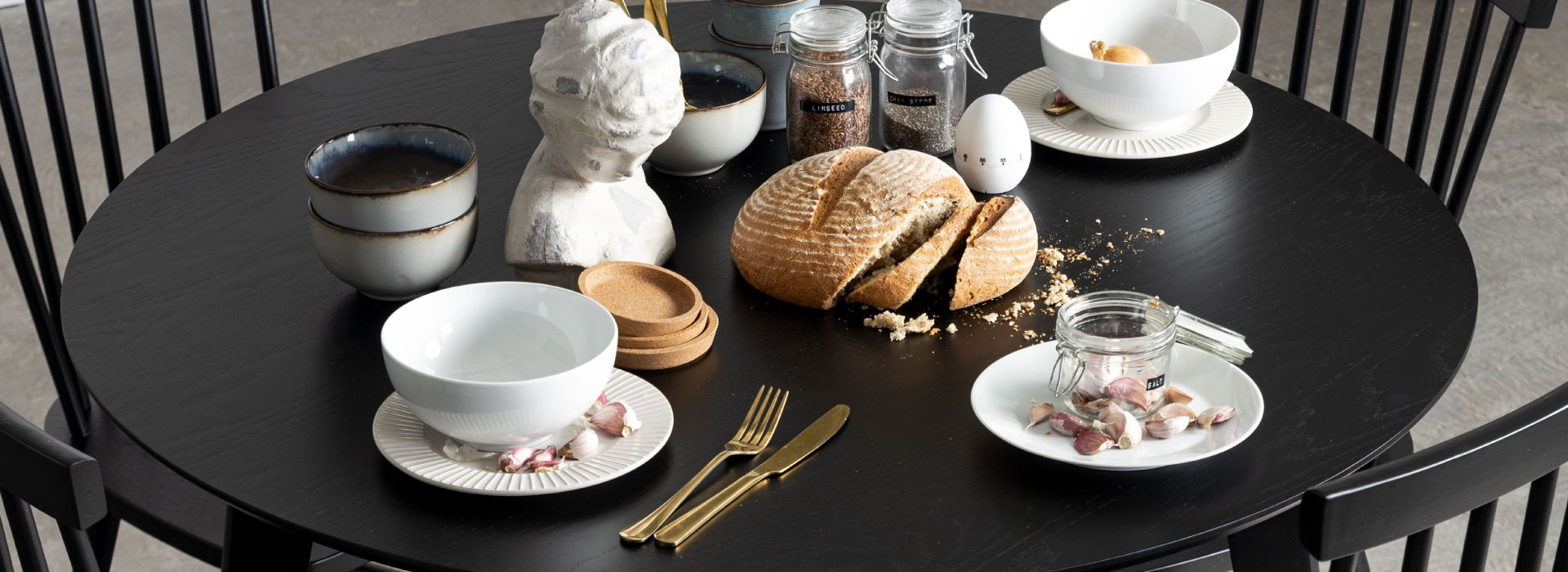 Zbliżenie na czarny stół do jadalni, na którym widać zastawę w kolorze błękitnym i białym, rozkrojony chleb, a także słoiki z musli i ziarnami.