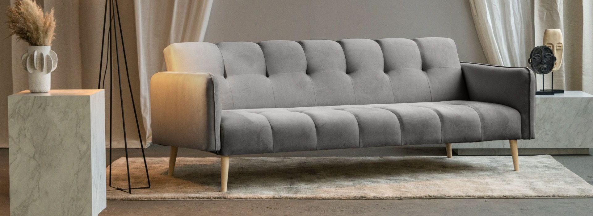 Szara sofa rozkładana na drewnianych nóżkach z dekoracyjnym zagłówkiem i siedziskiem. Po obu stronach sofy widoczne marmurowe stoliki.