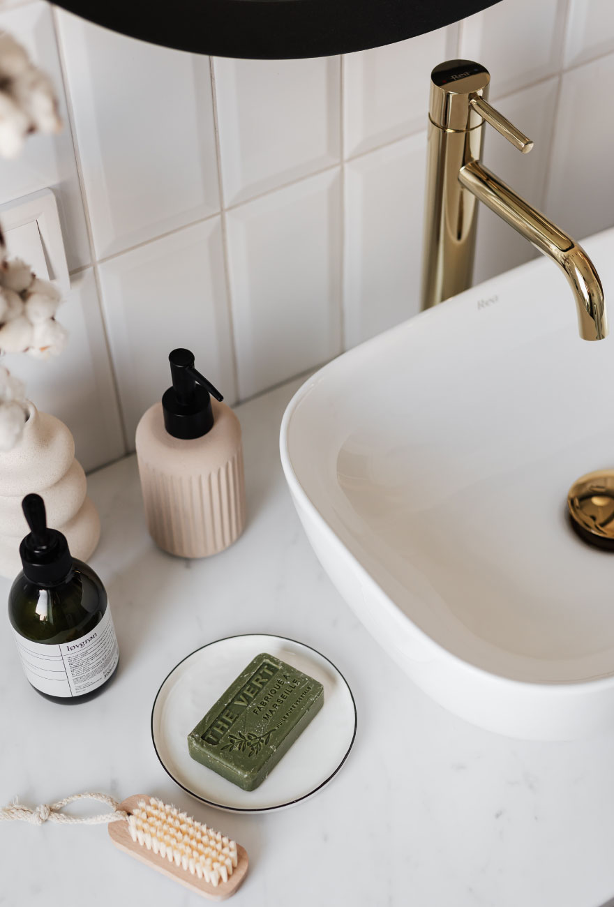 Zbliżenie na blat i umywalkę złotą armaturą w łazience. Wokół umywalki ułożone różne elementy, takie jak pojemnik na mydło, kosmetyki, szczoteczka do ciała oraz mydło.