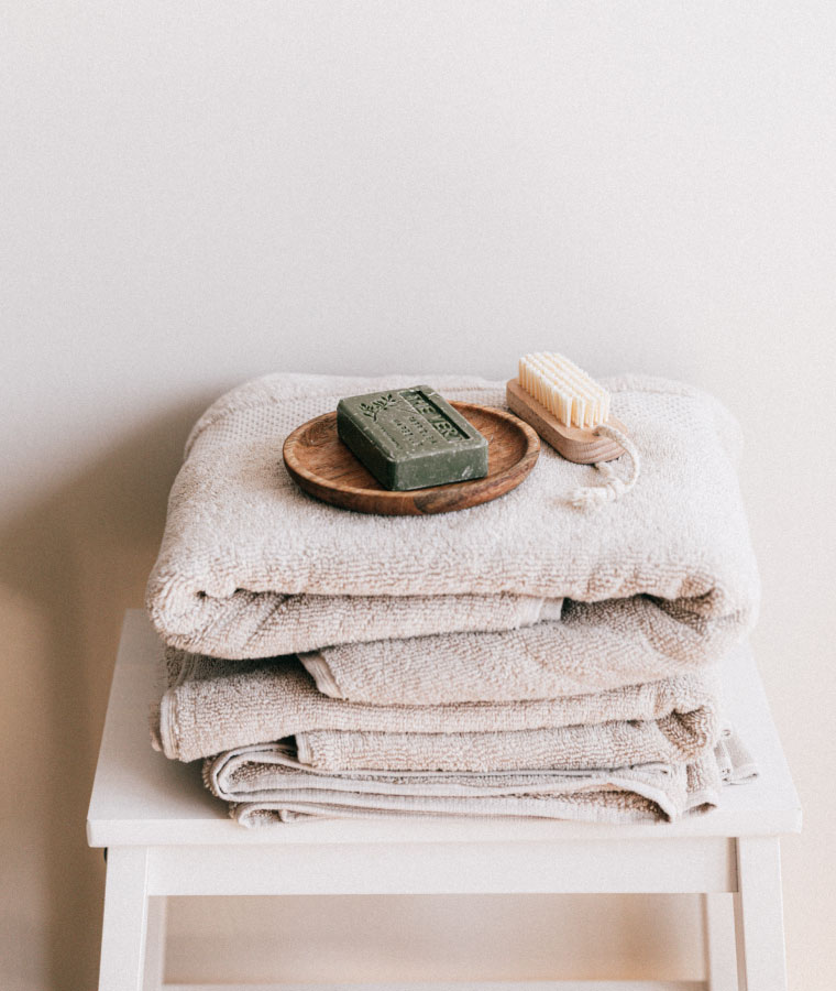 Drewniany biały taboret, na którym leżą złożone beżowe ręczniki, a na samej górze drewniana tacka z mydłem i szczoteczką do ciała.
