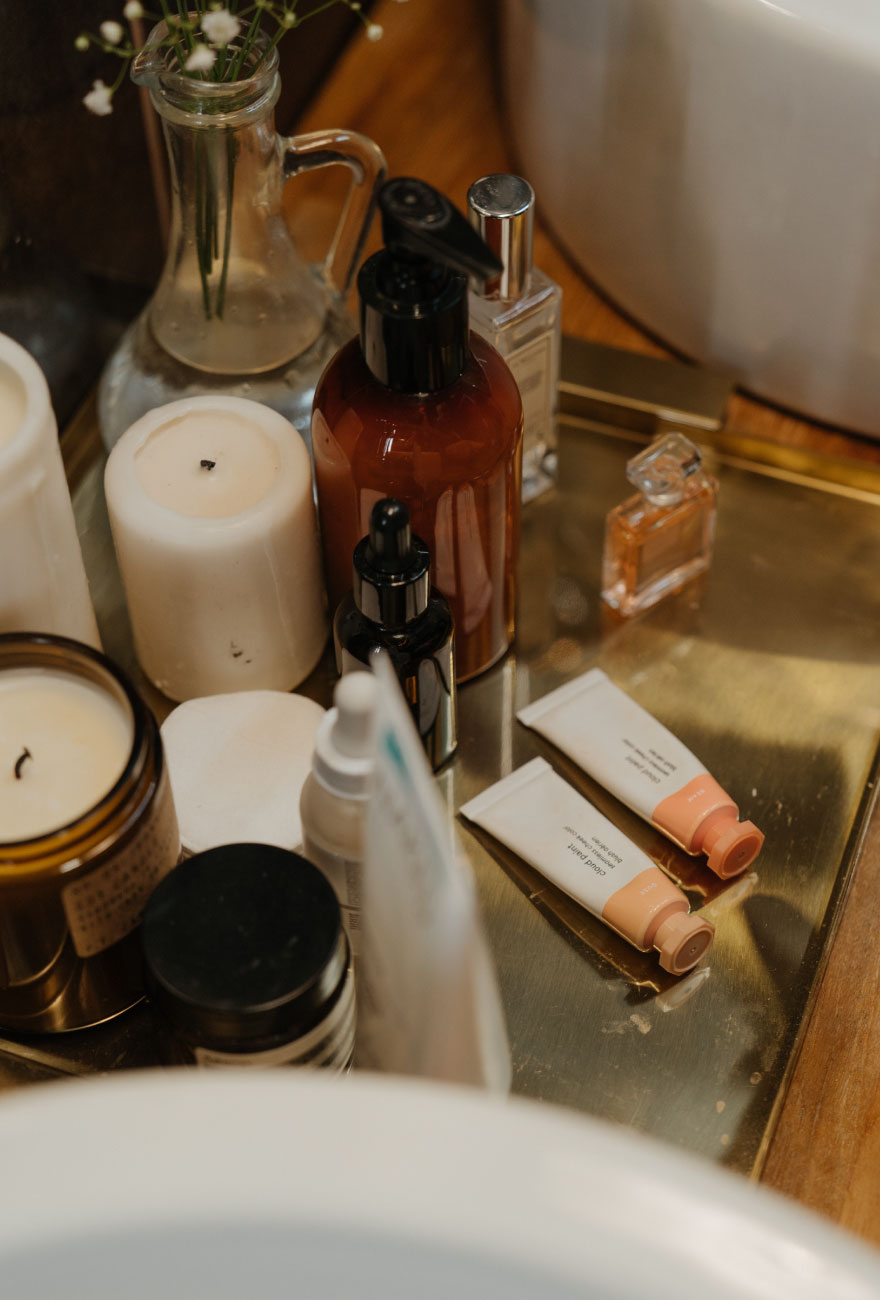 Zbliżenie na blat łazienki, na którym stoją różne kosmetyki, kremy, perfumy, a także świece w szklanych słoikach.