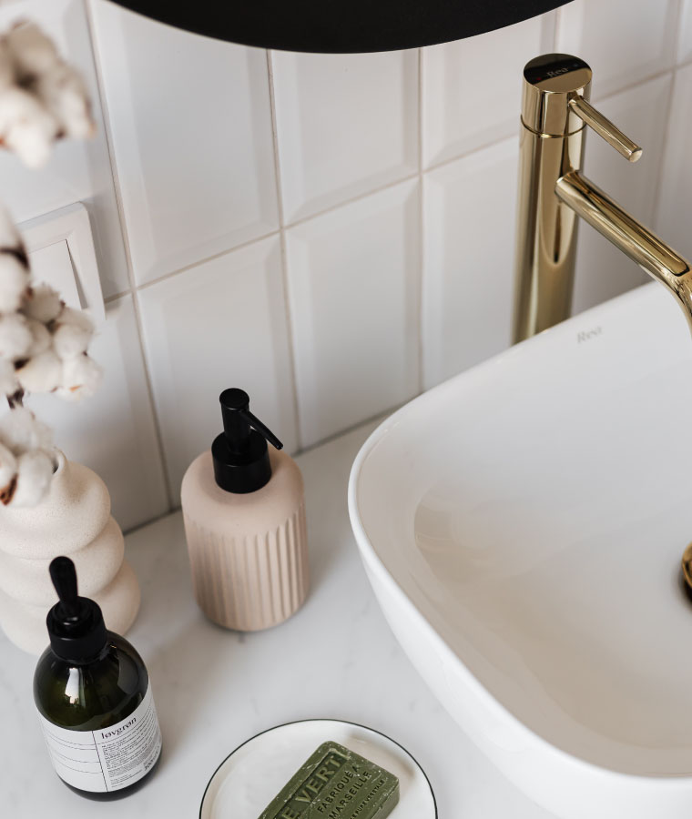 Zbliżenie na blat w łazience, na którym, obok umywalki, stoi dozownik do mydła, pojemnik na kosmetyki oraz mydło na dekoracyjnym talerzyku. W rogu zdjecia beżowy wazon z gałązką bawełny.