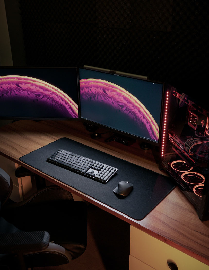 Wnętrze pokoju gamingowego z widocznymi dwoma monitorami, dużą, czarną podkładką pod klawiaturę i mysz oraz designerski komputer z otwartym wnętrzem.
