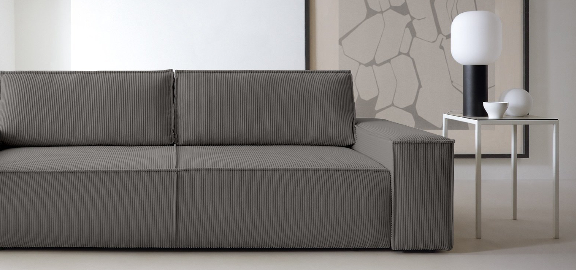 Ciemnoszara sofa w sztruksową tkaninę o bardzo prostej i geometrycznej formie. W tle duży, abstrakcyjny obraz, po prawej stronie sofy stolik pomocniczy oraz lampa stołowa na czarnej nodze.