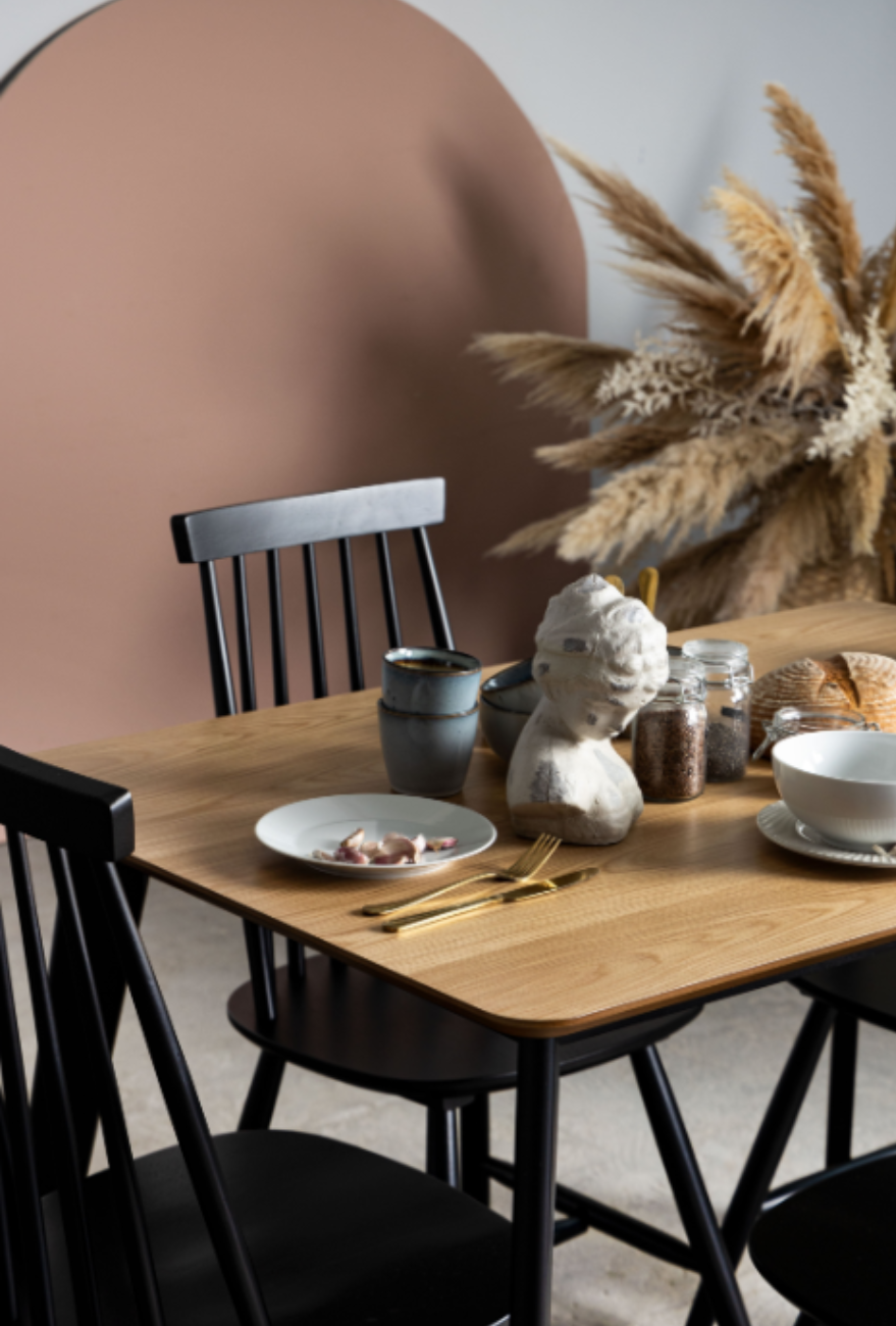 Drewniany stół i krzesła w czarnym kolorze. Blat stołu w kolorze jasnego drewna. Na stole ułożone naczynia oraz dekoracje. W tle trawy w donicy.