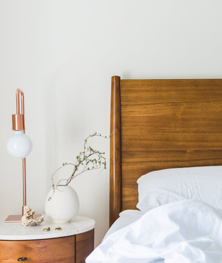 Zbliżenie na zagłówek łóżka wykonany z drewna, na łóżku biała pościel, a po lewej stronie stolik wykonany z drewna i marmuru.