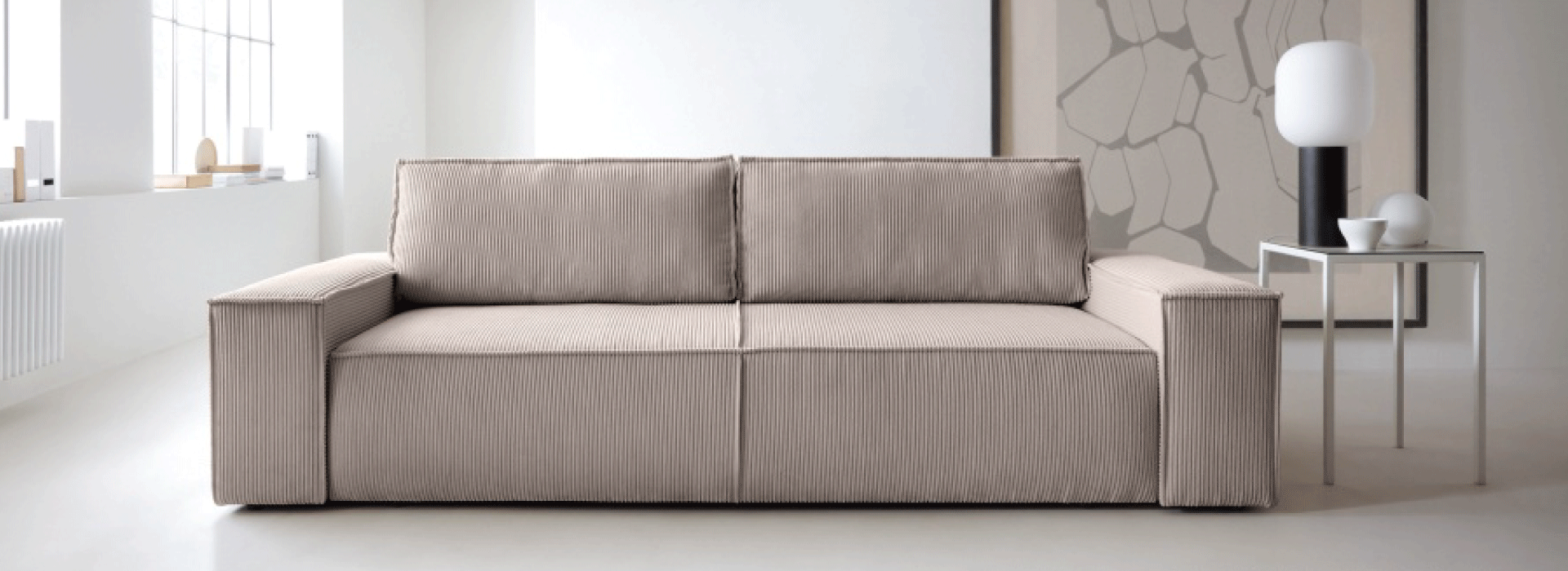 Beżowa sofa w minimalistycznym salonie z dużymi oknami. Na stoliku obok znajduje się lampa o nowoczesnym designie, a w tle widać abstrakcyjny obraz w neutralnych kolorach.