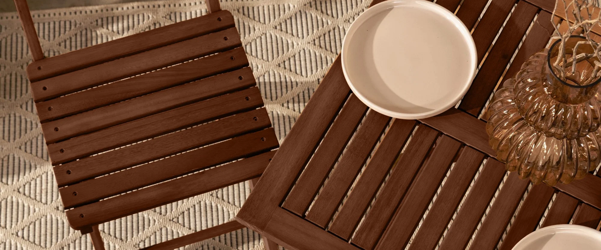 Drewniany stół i krzesło o ciemnobrązowym wykończeniu stoją na beżowym dywanie z geometrycznym wzorem. Na stole znajdują się jasna ceramiczna miseczka oraz dekoracyjny szklany wazon o żłobionej powierzchni z suchymi gałązkami.