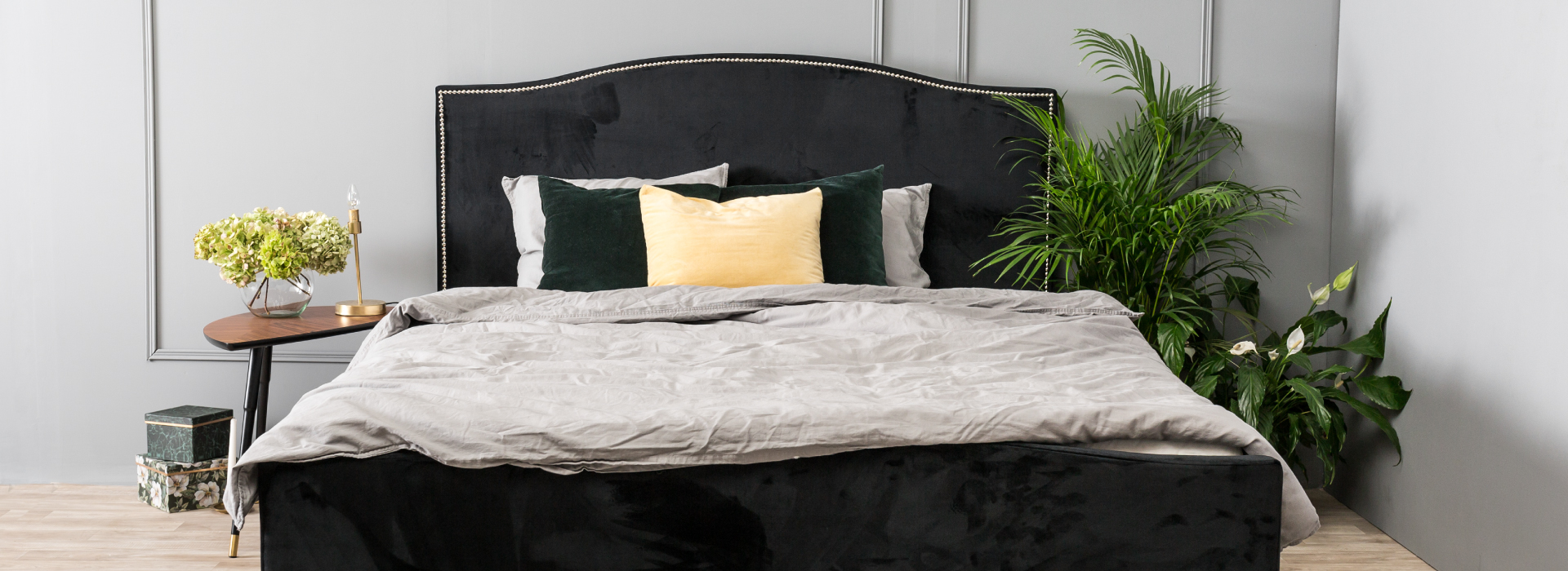 Granatowe łóżko obite welurową tkaniną ze zdobnym zagłówkiem, obok łóżka rośliny, a po drugiej stronie drewniany stolik.