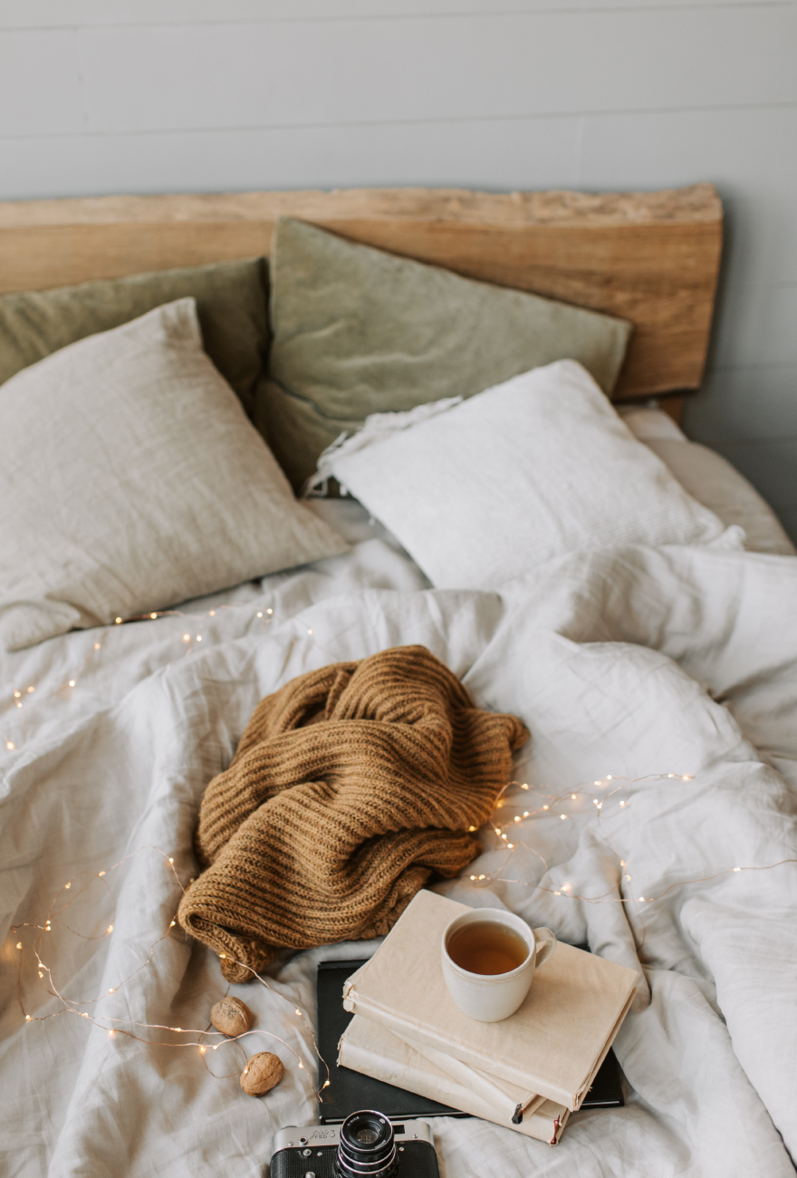 Łóżko z drewnianym zagłówkiem, biało-zieloną pościelą oraz stertą książek z kubkiem herbaty leżącymi na nim, obok karmelowy koc i aparat fotograficzny.