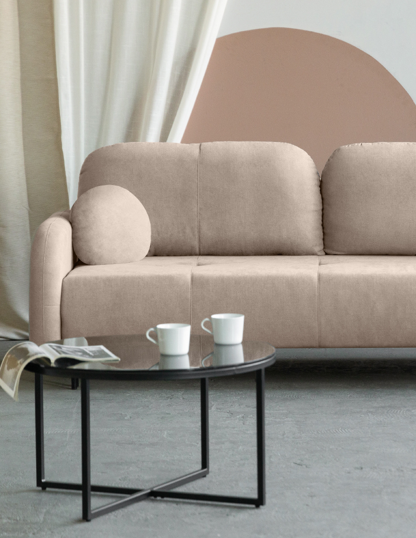 Beżowa sofa z zaokrąlonymi poduszkami oparciowymi i jedną mniejszą okrągłą poduszką po lewej stronie. Przed sofą czarny stolik kawowy z gazetą i dwoma kubkami.