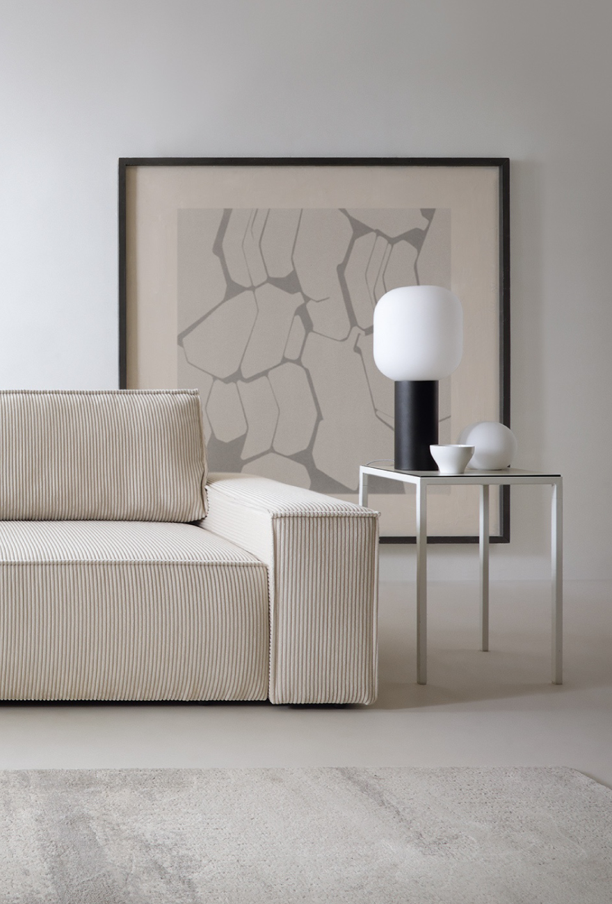 Jasnobeżowa sofa w tkaninie sztruksowej w organicznym, kanciastym kształcie. Po prawej stronie stolik kawowy z dekoracjami, w tym lampką na czarnej podstawie.