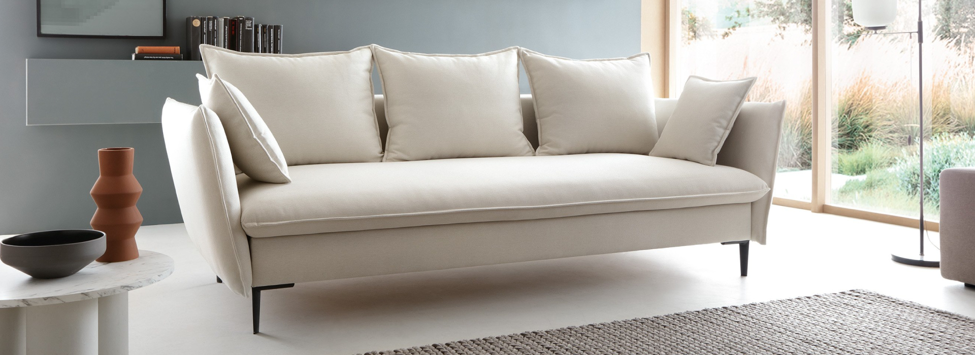 Jasnobeżowa sofa z trzema dużymi poduszkami i dwoma mniejszymi na czarnych, metalowych nóżkach. W tle ściana z półką oraz biały stolik kawowy.