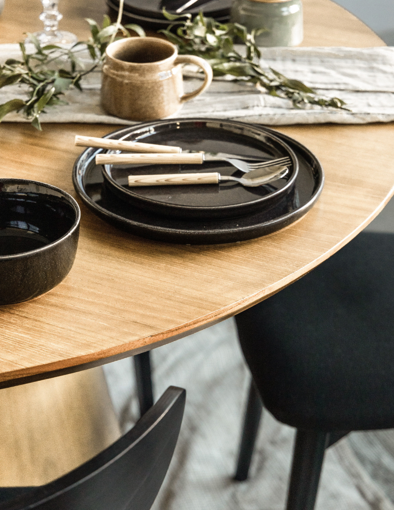 Czarne krzesła z materiałowymi siedziskami stojąca wokół okrągłego drewnianego stołu. Na stole zastawa w kolorze czarnym i naturalne dekoracje.