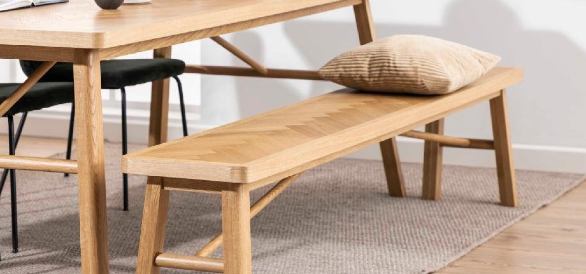 Drewniana ławka stojąca wzdłuż długiego boku drewnianego stołu. Na ławce beżowa poduszka w drobne paski.