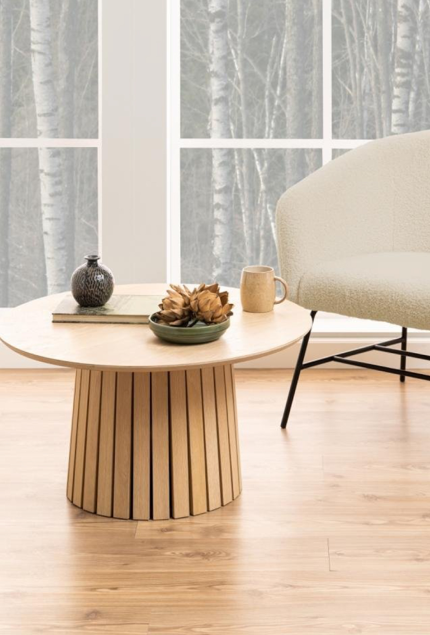 Drewniany stolik z lamelową podstawą i okrągłym blatem, na którym stoją jesienne dekoracje. Obok stolika fotel w tkaninie boucle na czarnej konstrukcji.